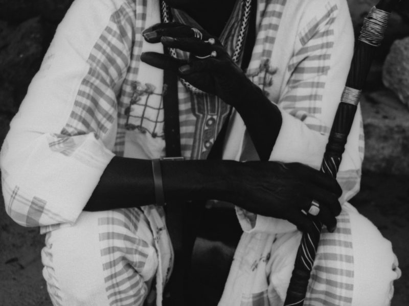 Black and white photo of a person in a patterned robe holding a staff. The focus is on their hands and the fabric, with intricate details of rings and bracelets visible.