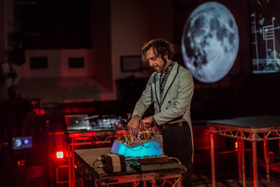 A man in a suit meticulously operates a complex electronic device with wires sprawled across the table. Behind him, a large, illuminated moon is projected on a screen