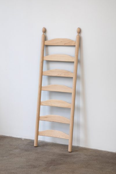 A light wooden decorative ladder with six curved rungs leans against a white wall on a beige carpeted floor.