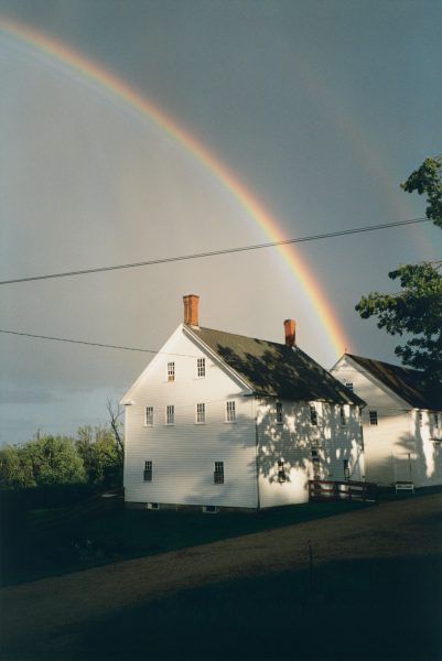 A charming white house with a pitched roof stands under a vibrant rainbow in a rural setting. Soft shadows are cast on the building, and a trees branches frame part of the view. The sky is overcast, adding a serene ambiance.