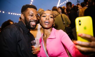 A man in a black shirt and a woman in a pink outfit snap a selfie as string lights twinkle behind them.