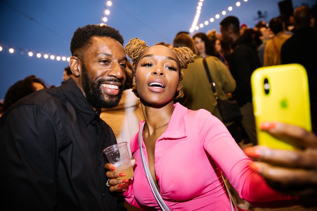 A man in a black shirt and a woman in a pink outfit snap a selfie as string lights twinkle behind them.