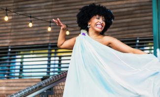 A joyful person with curly hair is smiling and posing with one arm raised, wearing a flowing light blue dress on an outdoor stairway adorned with string lights.