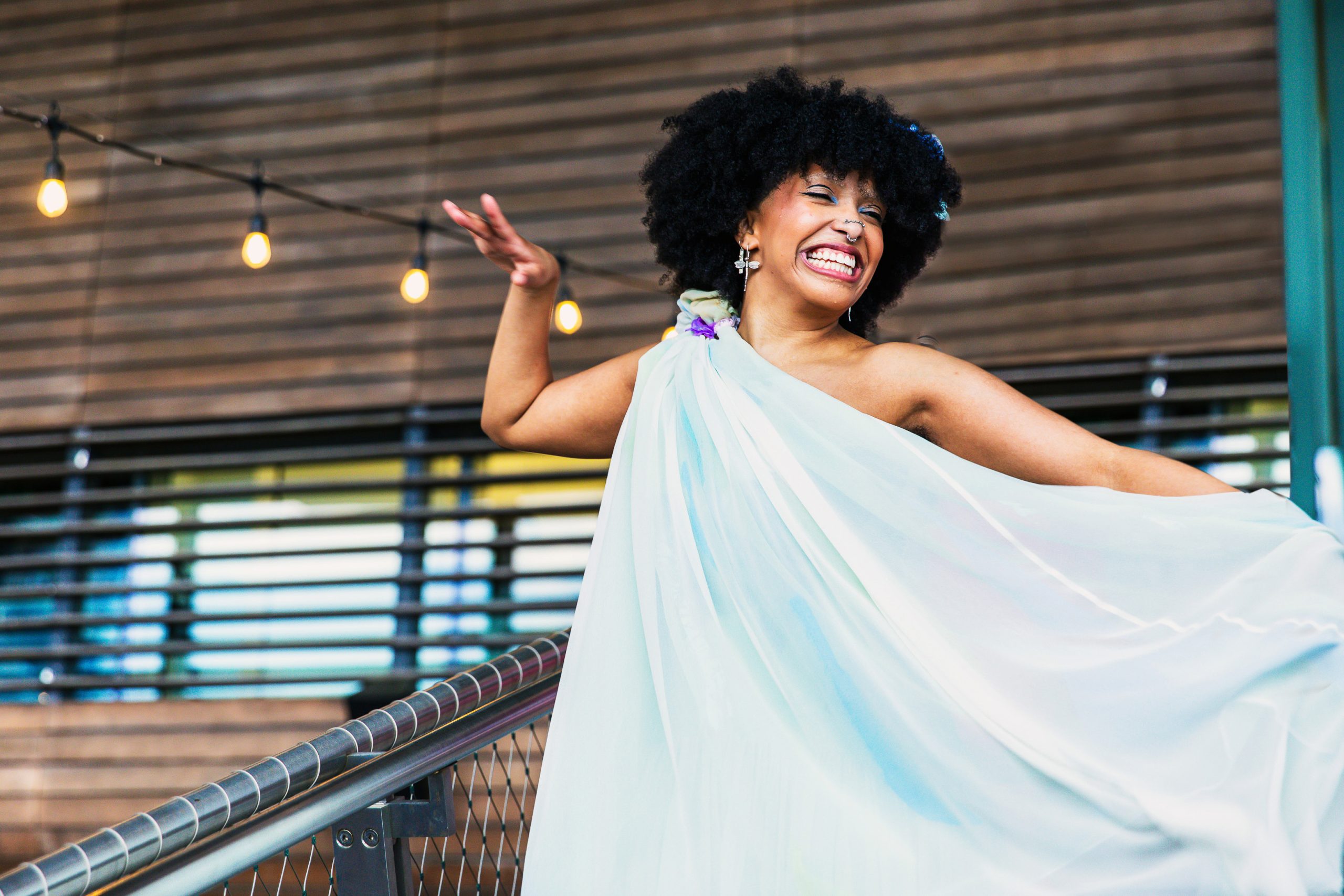 A joyful person with curly hair is smiling and posing with one arm raised, wearing a flowing light blue dress on an outdoor stairway adorned with string lights.