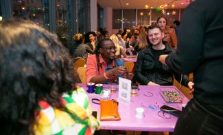 People smiling at tables while making art with beads and threads and enjoying drinks. A person in the foreground hands an item to another while others engage in conversation.