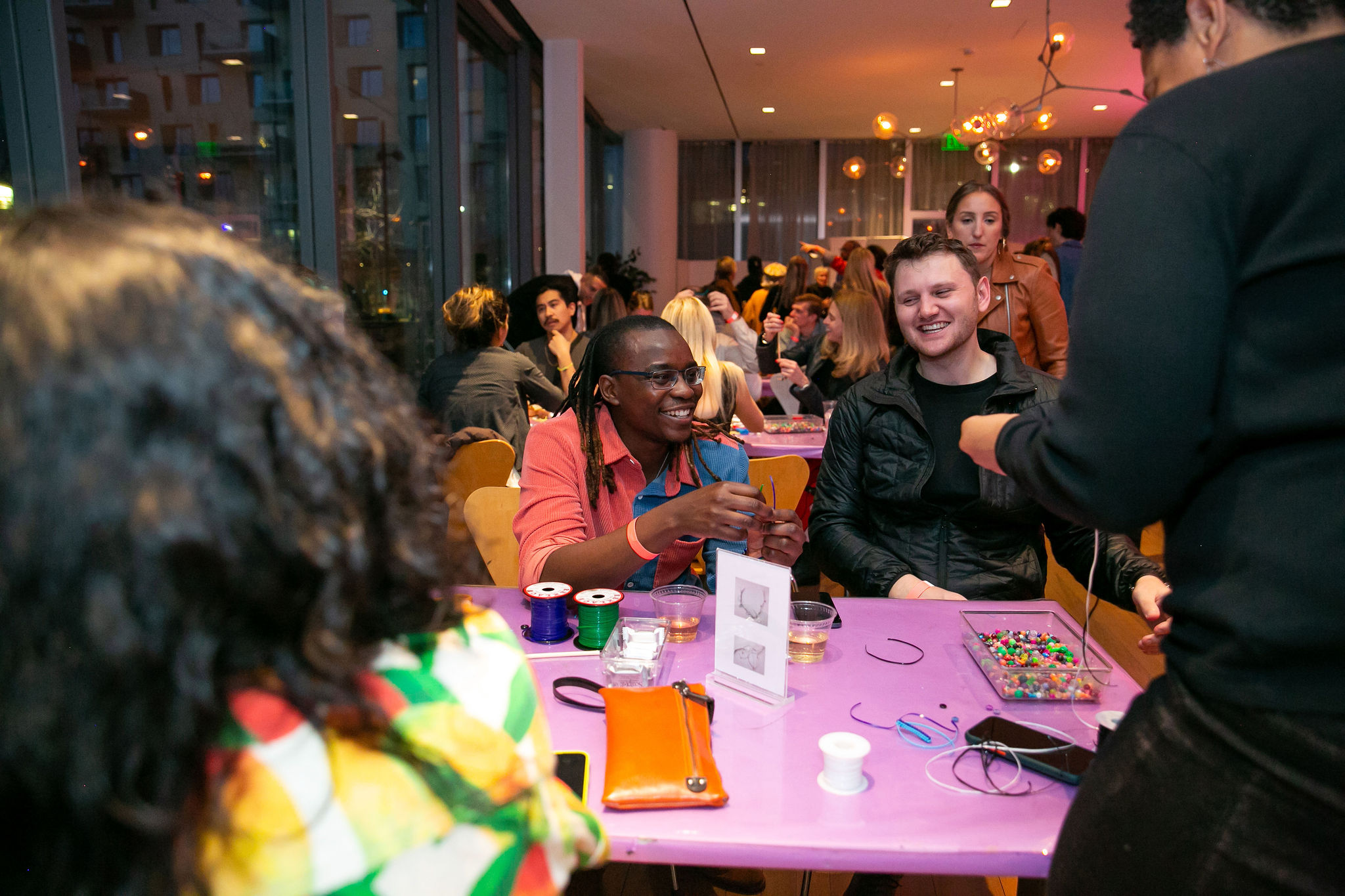 People smiling at tables while making art with beads and threads and enjoying drinks. A person in the foreground hands an item to another while others engage in conversation.