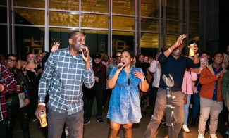 A lively indoor gathering at Boots and Buckles with three people singing passionately into microphones. The First Fridays crowd around them is cheering and reveling in the music. The venue boasts large windows and ambient lighting, creating an inviting atmosphere.