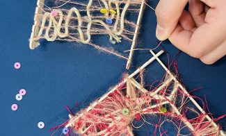 Two hands weave yarn and colorful beads onto small square looms made of wooden sticks against a blue background. Loose beads and unfinished weavings are scattered around, showcasing a crafting activity.