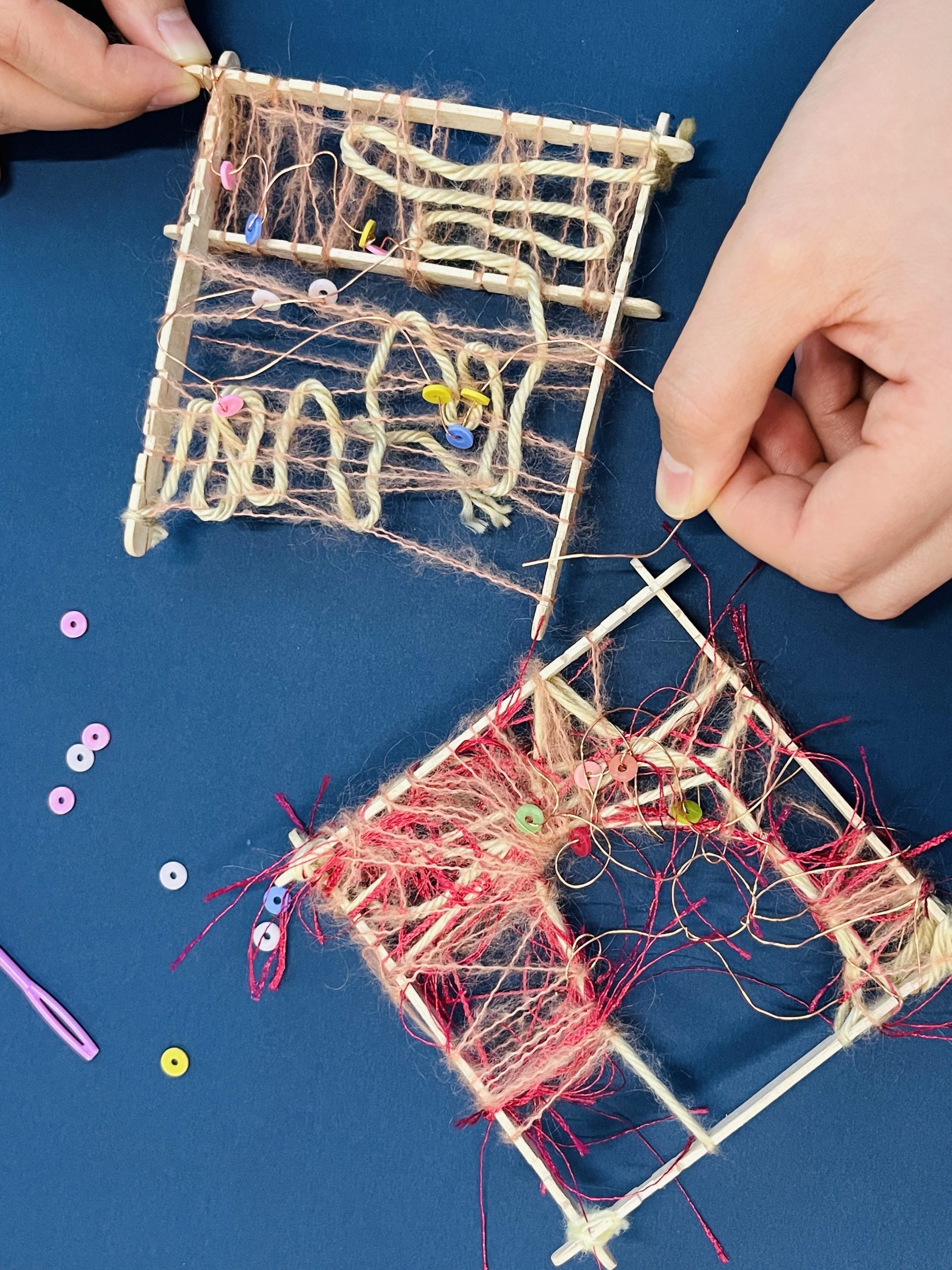 Two hands weave yarn and colorful beads onto small square looms made of wooden sticks against a blue background. Loose beads and unfinished weavings are scattered around, showcasing a crafting activity.