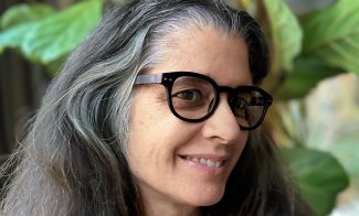 A woman with long gray hair and black glasses smiles, wearing a dark blue top. She stands against lush green plants