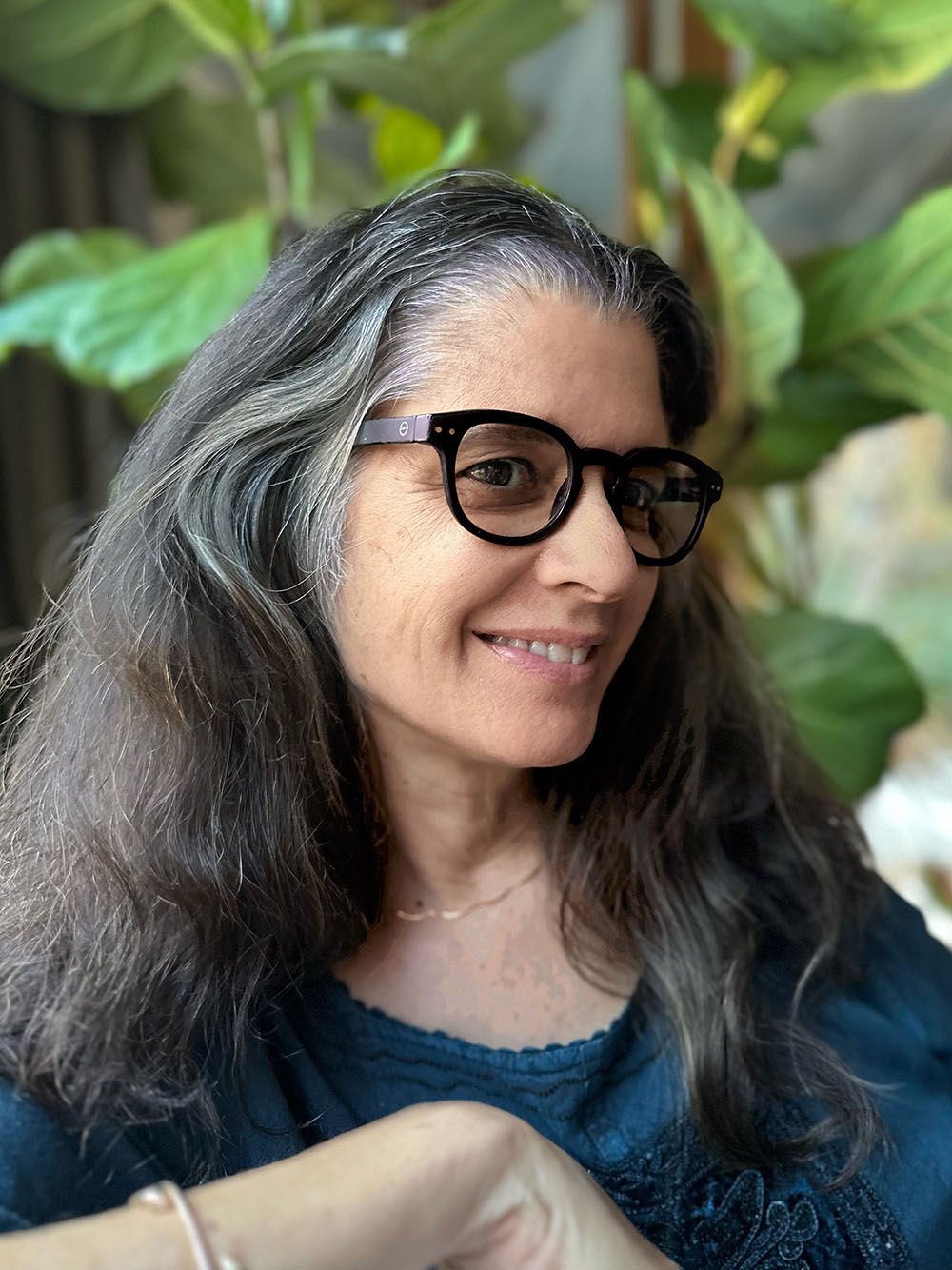 A woman with long gray hair and black glasses smiles, wearing a dark blue top. She stands against lush green plants