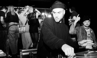 A DJ in a beanie is focused on mixing music at a turntable in a lively club setting