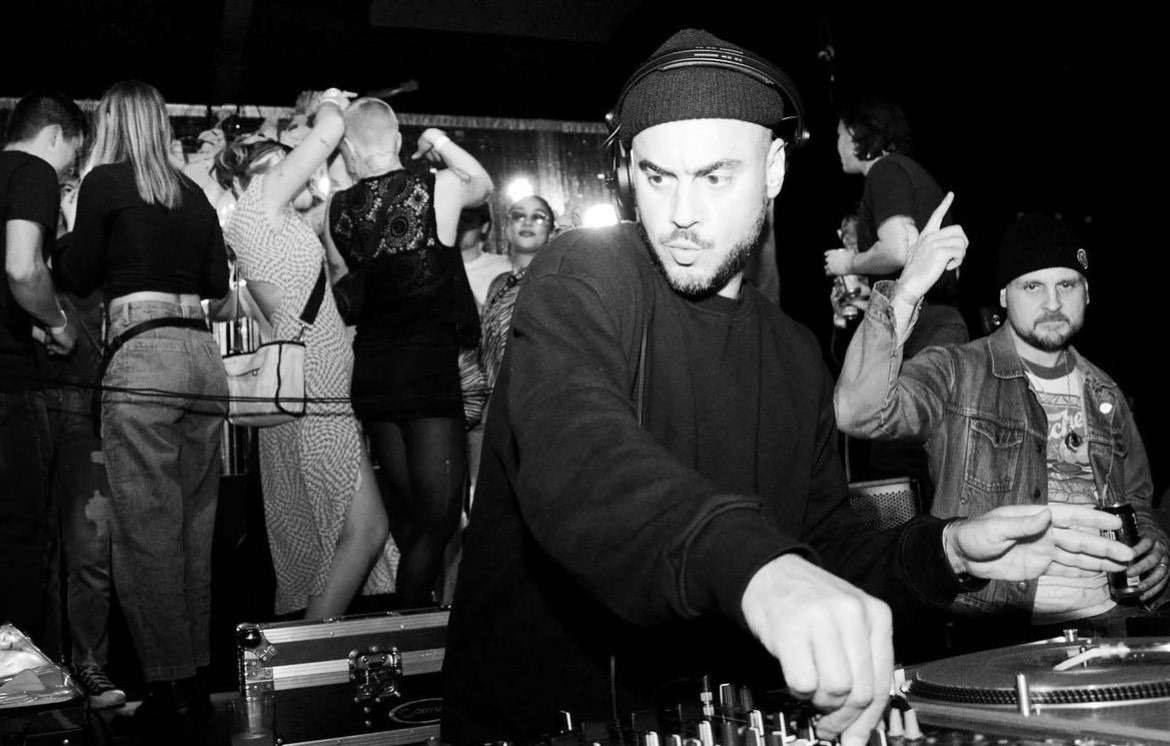 A DJ in a beanie is focused on mixing music at a turntable in a lively club setting