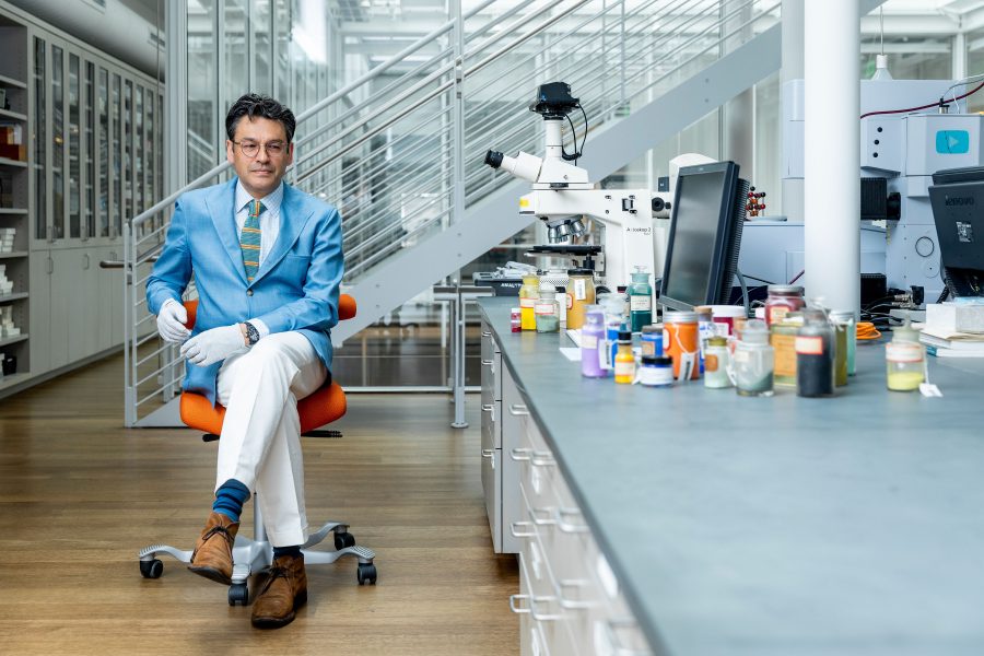 A person in a blue blazer and white pants sits on an orange chair in a modern laboratory, exuding the precision of an artist. They are wearing glasses and gloves. The workspace, reminiscent of The Artist's Voice, features microscopes and various jars on the countertop. A staircase is visible in the background.