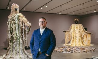 A man in glasses and a blue jacket stands in an art gallery. Behind him are two large, elaborate sculptures wearing flowing garments, each with unique textures and colors