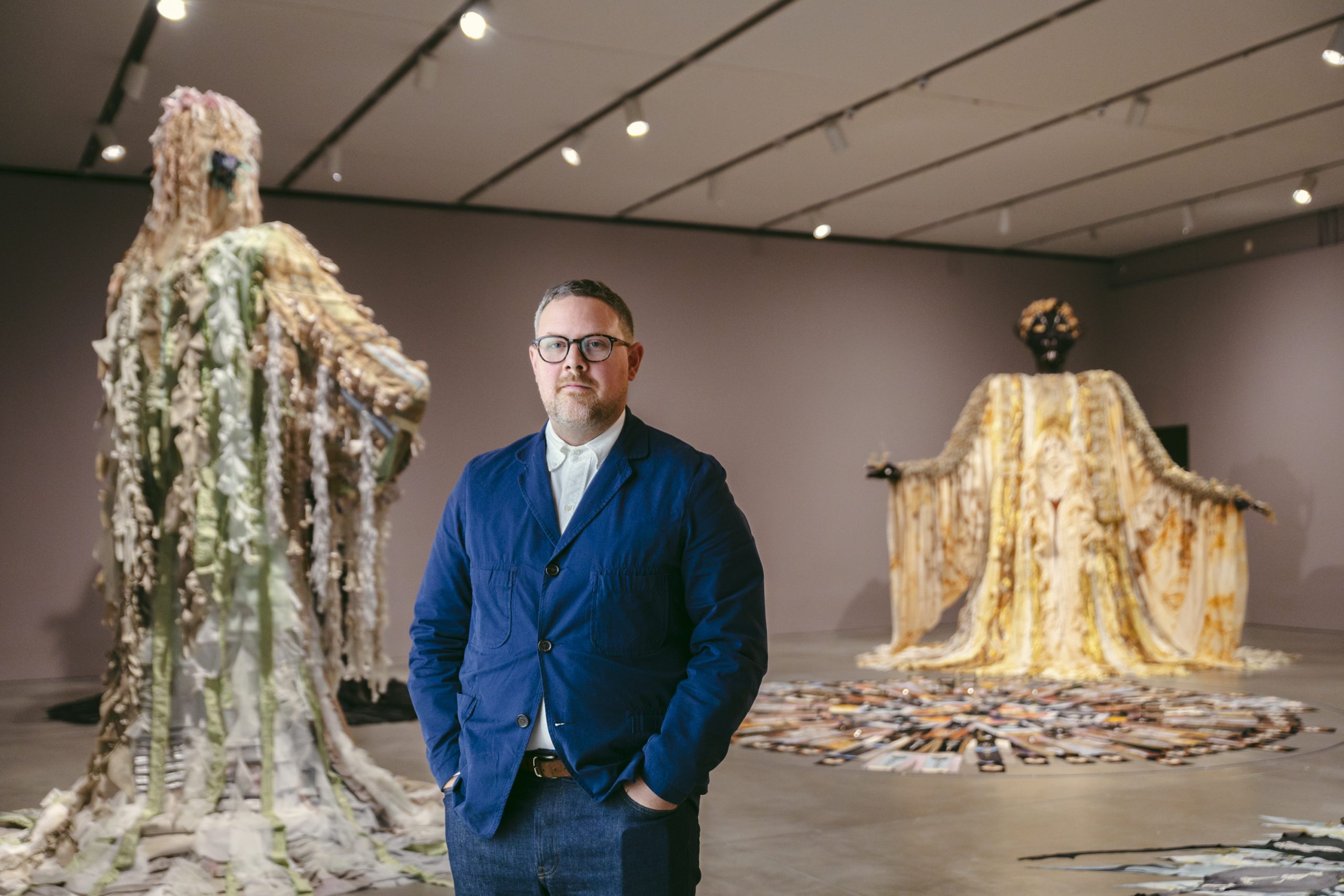 A man in glasses and a blue jacket stands in an art gallery. Behind him are two large, elaborate sculptures wearing flowing garments, each with unique textures and colors