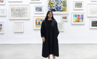 A woman in a long, black dress stands in front of a white wall covered with colorful, framed abstract artwork