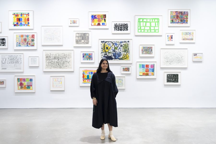 A woman in a long, black dress stands in front of a white wall covered with colorful, framed abstract artwork at the ICA Boston, just before Tessa Bachi Haas begins her gallery talk.