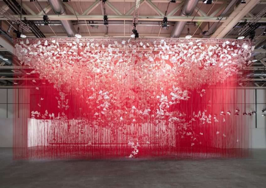 A mesmerizing art installation features countless white paper butterflies suspended in the air, creating an illusion of fluttering motion. They hang from red strings, forming a flowing, cloud-like formation against an industrial-style ceiling.