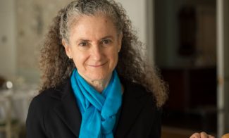 A woman with curly gray hair smiles at the camera. She is wearing a bright blue scarf and a dark jacket. The background is softly blurred, featuring light-colored walls and furniture.