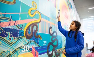 A girl in a blue hoodie with braided hair stands before a colorful mural featuring a snake motif and numbered squares. She holds up a large metal piece