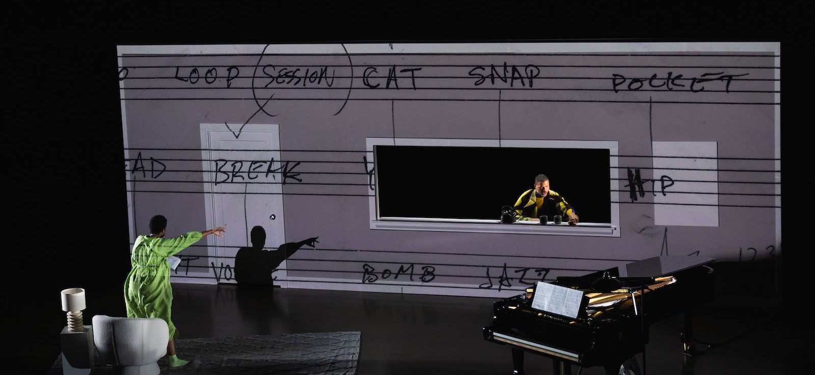 A performer in a green outfit gestures toward a large, illuminated screen with musical notations. Another figure in yellow is visible through a window in the screen. A grand piano and a small round table with a lamp are on stage.