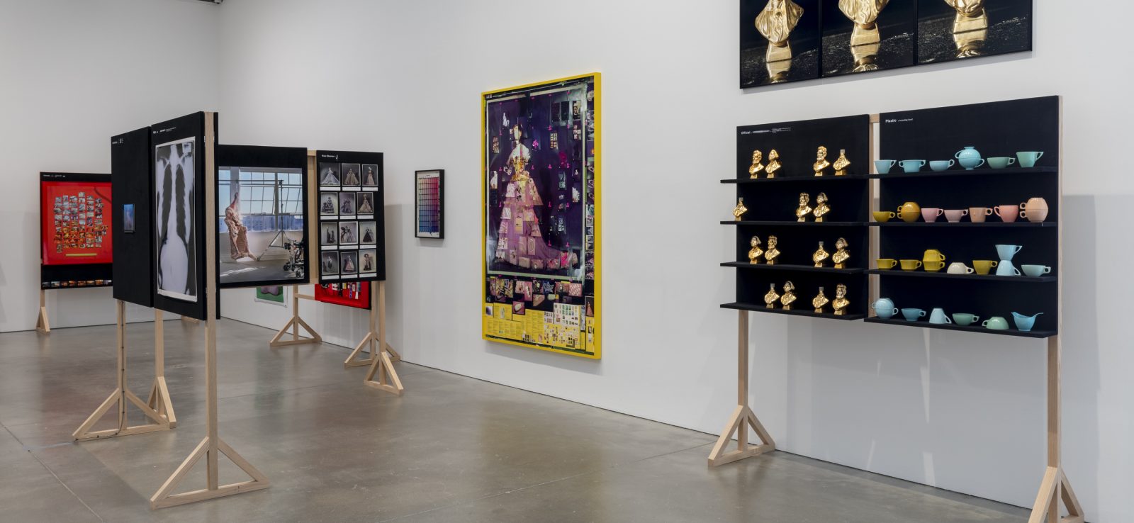 A gallery displaying various artworks. On the right, a black stand holds gold figurines and pastel bowls. Framed works and photographs are mounted on black panels on wooden frames.