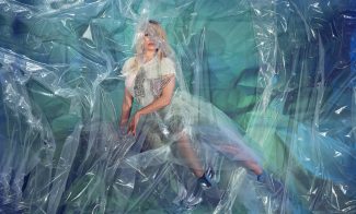 Pamela Anderson poses dramatically behind a transparent plastic sheet while wearing a graphic T-shirt and a tulle skirt