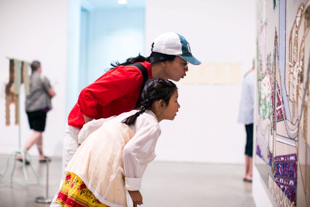 A woman in a red shirt and a white and blue cap leans over with a young girl in a white dress and yellow skirt. They closely observe a detailed artwork in the brightly lit gallery space, while other visitors are visible in the background.