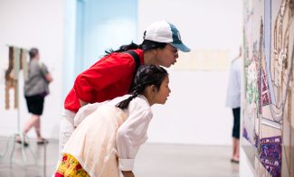 A woman in a red shirt and a white and blue cap leans over with a young girl in a white dress and yellow skirt. They closely observe a detailed artwork in the brightly lit gallery space, while other visitors are visible in the background.