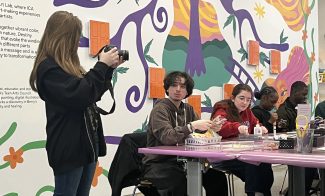 A person with long hair stands taking photos of a group of people sitting at a table. The wall behind them features colorful abstract artwork with a tree and various shapes. The people are engaged in crafts or artwork on the table.