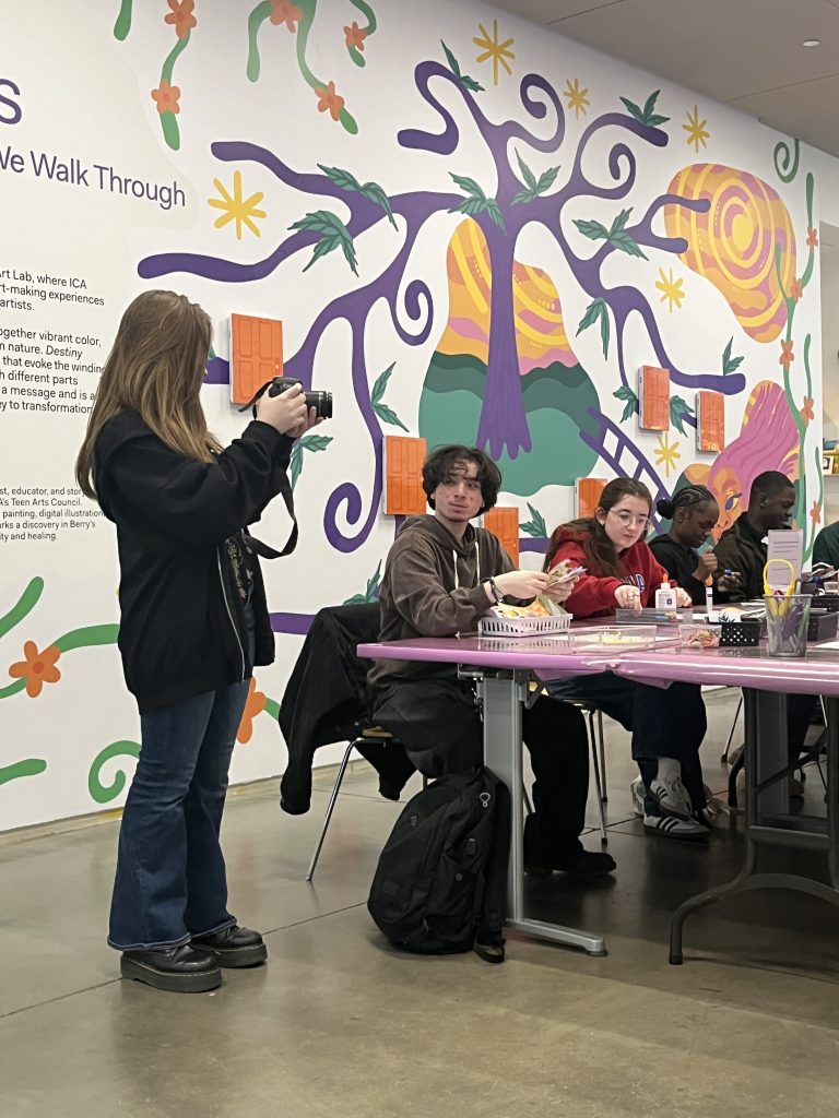 A person with long hair stands taking photos of a group of people sitting at a table. The wall behind them features colorful abstract artwork with a tree and various shapes. The people are engaged in crafts or artwork on the table.