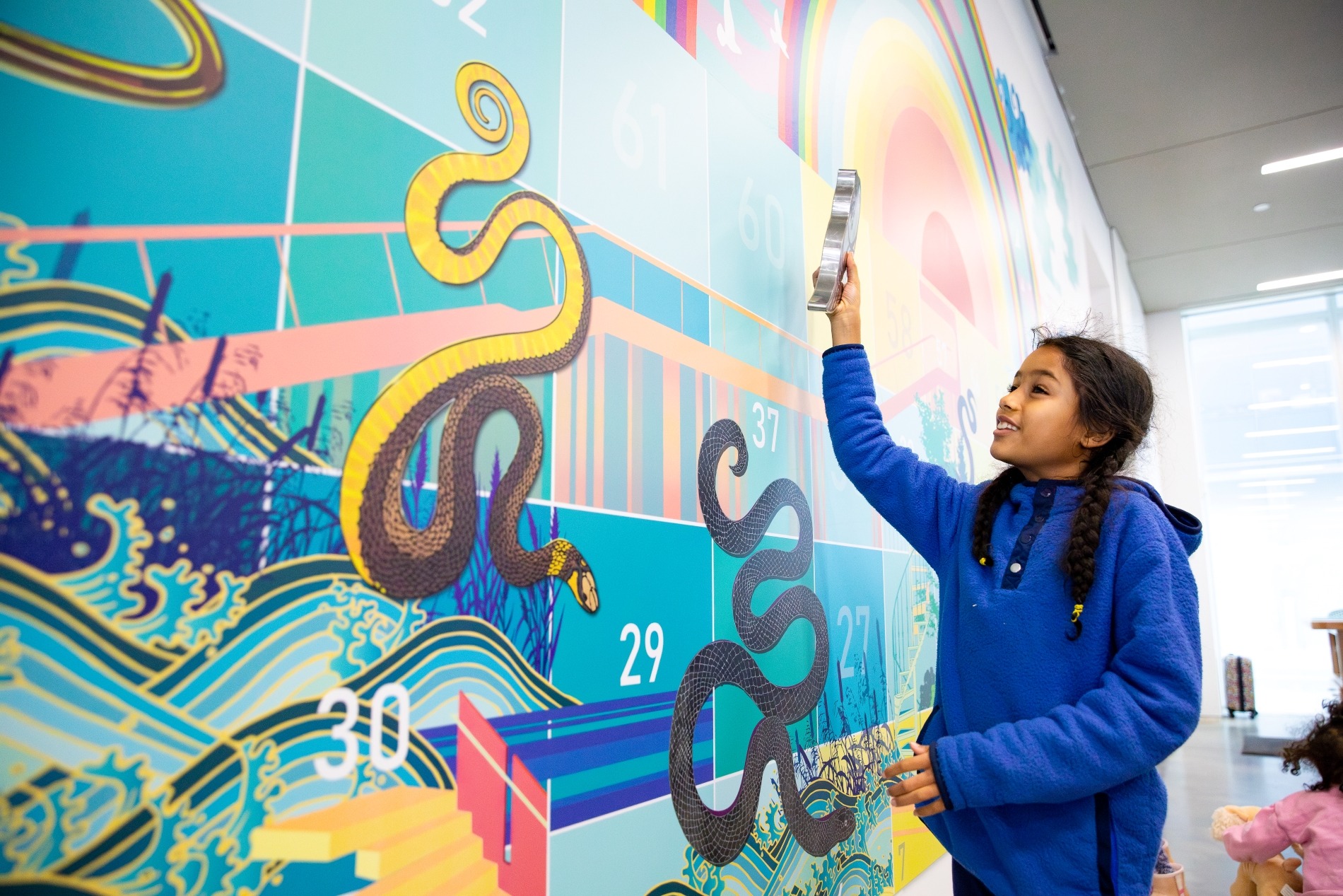 A young girl in a blue hoodie touches a colorful wall mural featuring snakes and numbers in a modern indoor space. Shes smiling and has long braided hair. Another child is partially visible to the left.