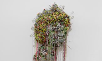 A vibrant, textured wall sculpture by Hew Locke composed of colorful plastic flowers and cascading strings of beads, it forms an abstract shape reminiscent of a head in profile