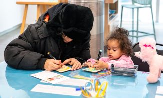 An adult and a child sit at a blue table, engaging in a crafting activity. The adult wears a black winter coat and hat, while the child, in a pink jacket, holds a marker. Nearby, there are a stuffed toy and various craft supplies.