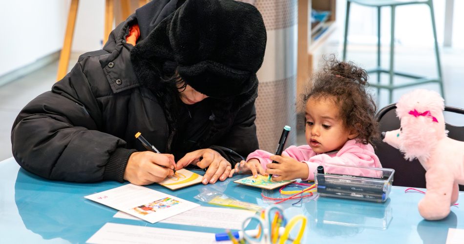 An adult and a child sit at a blue table, engaging in a crafting activity. The adult wears a black winter coat and hat, while the child, in a pink jacket, holds a marker. Nearby, there are a stuffed toy and various craft supplies.
