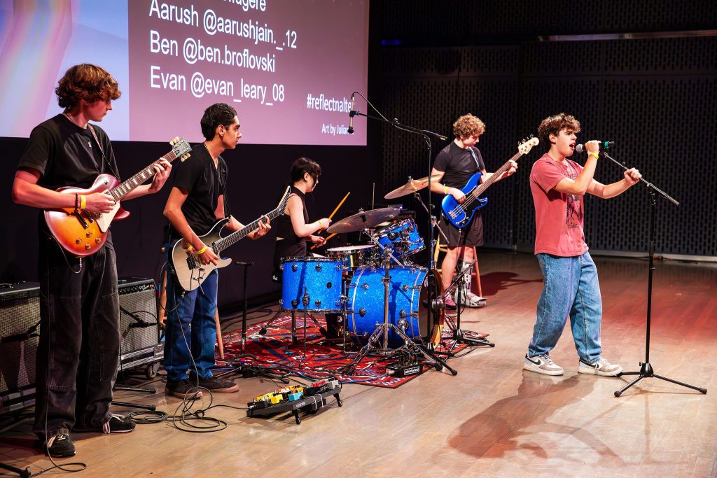 A band of five young musicians performs on stage. The drummer is seated behind a blue drum set, while two guitarists, a bassist, and a vocalist stand. A screen in the background displays social media handles. The stage is lit with colorful lights.