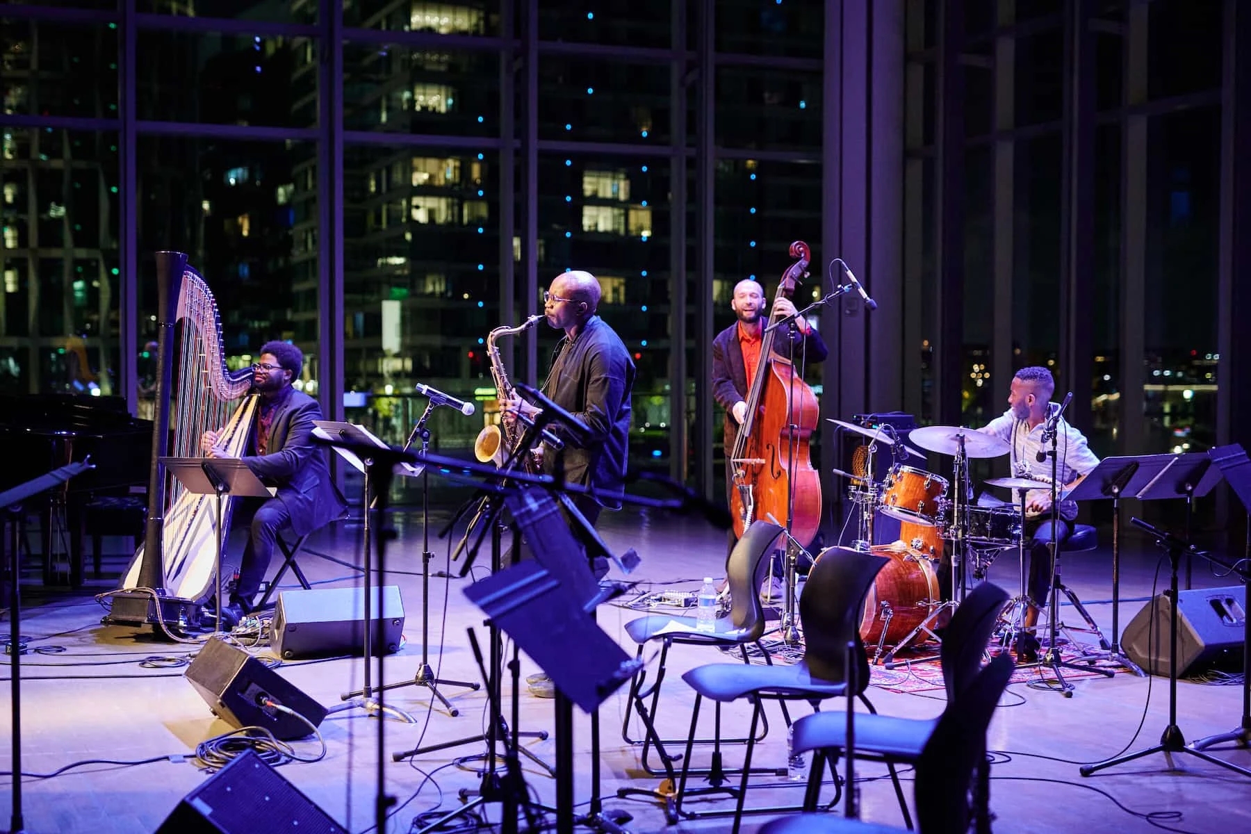 A jazz band performs on stage at the urbane ICA Boston, with a harpist, saxophonist, double bassist, and drummer. Inside this modern venue, large glass windows reveal a cityscape of lit buildings at night. Chairs and music stands are visible in the foreground, capturing the chic vibe of a hidden cafe.