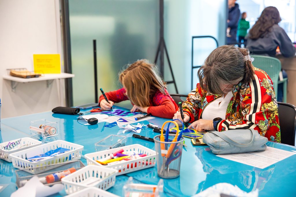 A woman and child sit at a blue table in a bright room, immersed in drawing with markers. The table overflows with paper, markers, and craft supplies. Other people are visible in the background