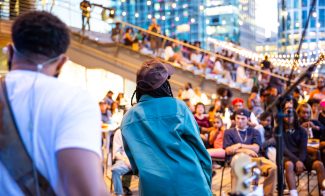 A performer in a teal jacket and brown cap faces an audience seated outdoors in an urban area at dusk. String lights hang above, and city buildings reflect in the background.