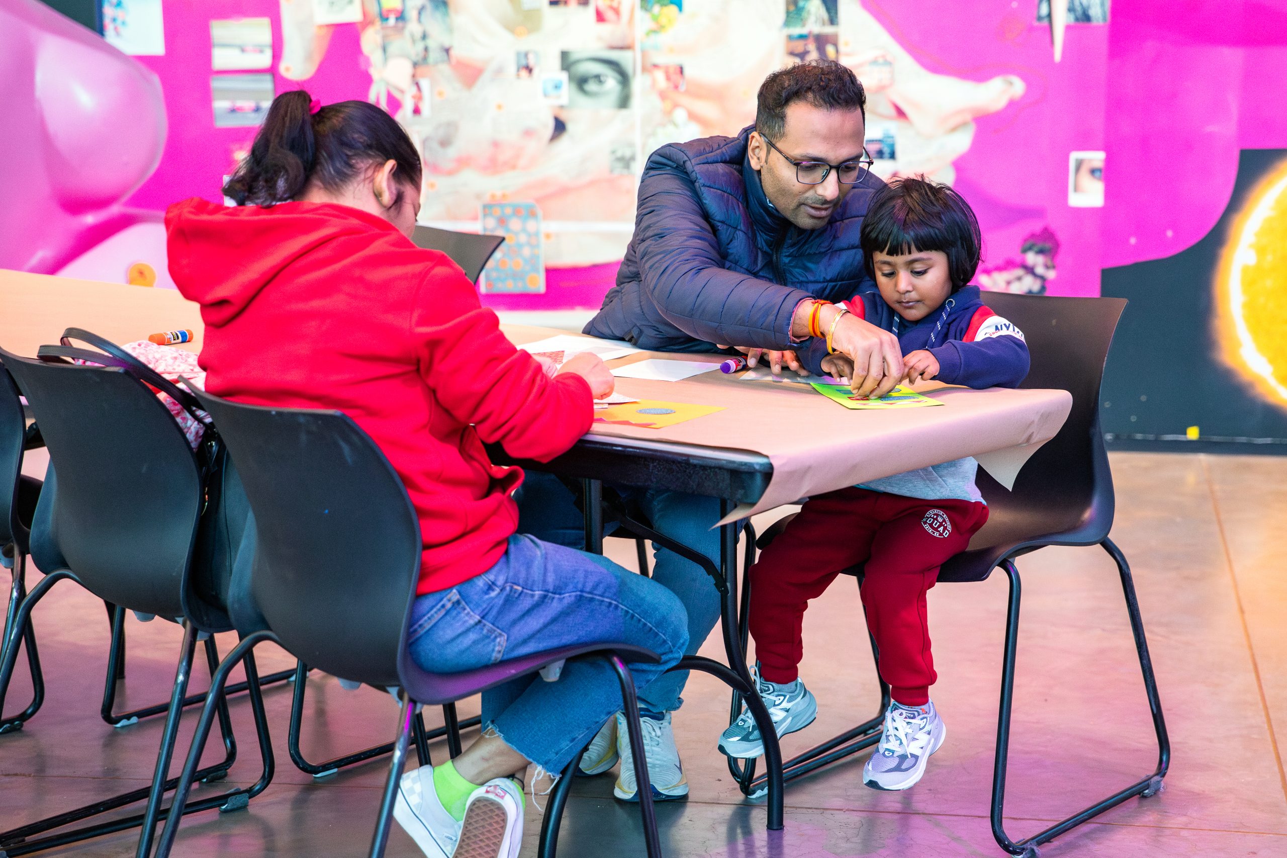 a child and a man are crafting at a table covered, in front of a bright mural. The man assists the child, both focused on their activity, while another person in a red hoodie sits nearby.