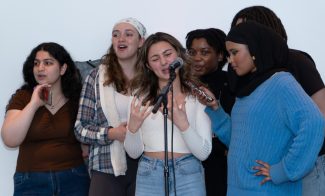 A group of six young people stand together, one holding a microphone and others holding phones, as one person in the center passionately sings or speaks into a microphone. They appear engaged and expressive.