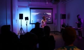 A performer with a microphone stands in front of a projected screen showing a car. Audience members watch in a dimly lit room with blue and purple lighting. Music equipment is visible on the right.