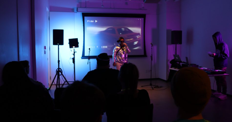 A performer with a microphone stands in front of a projected screen showing a car. Audience members watch in a dimly lit room with blue and purple lighting. Music equipment is visible on the right.