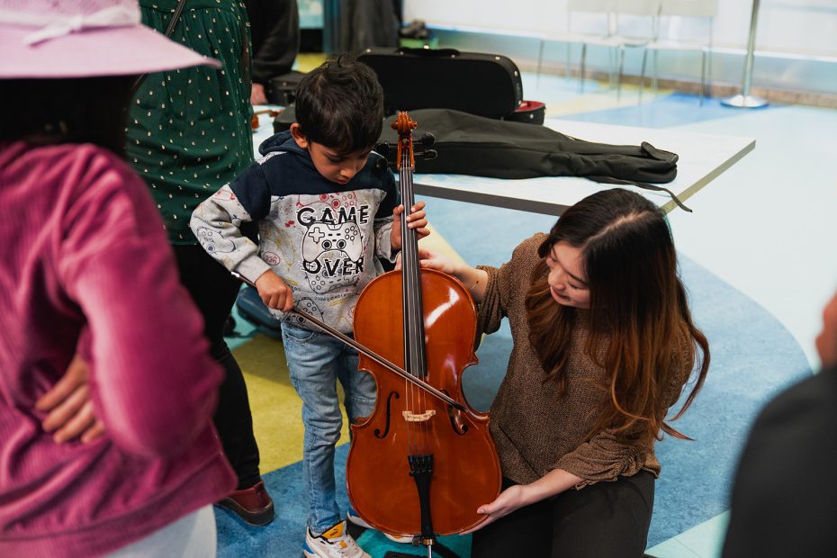 A young child plays a cello, with guidance from a woman