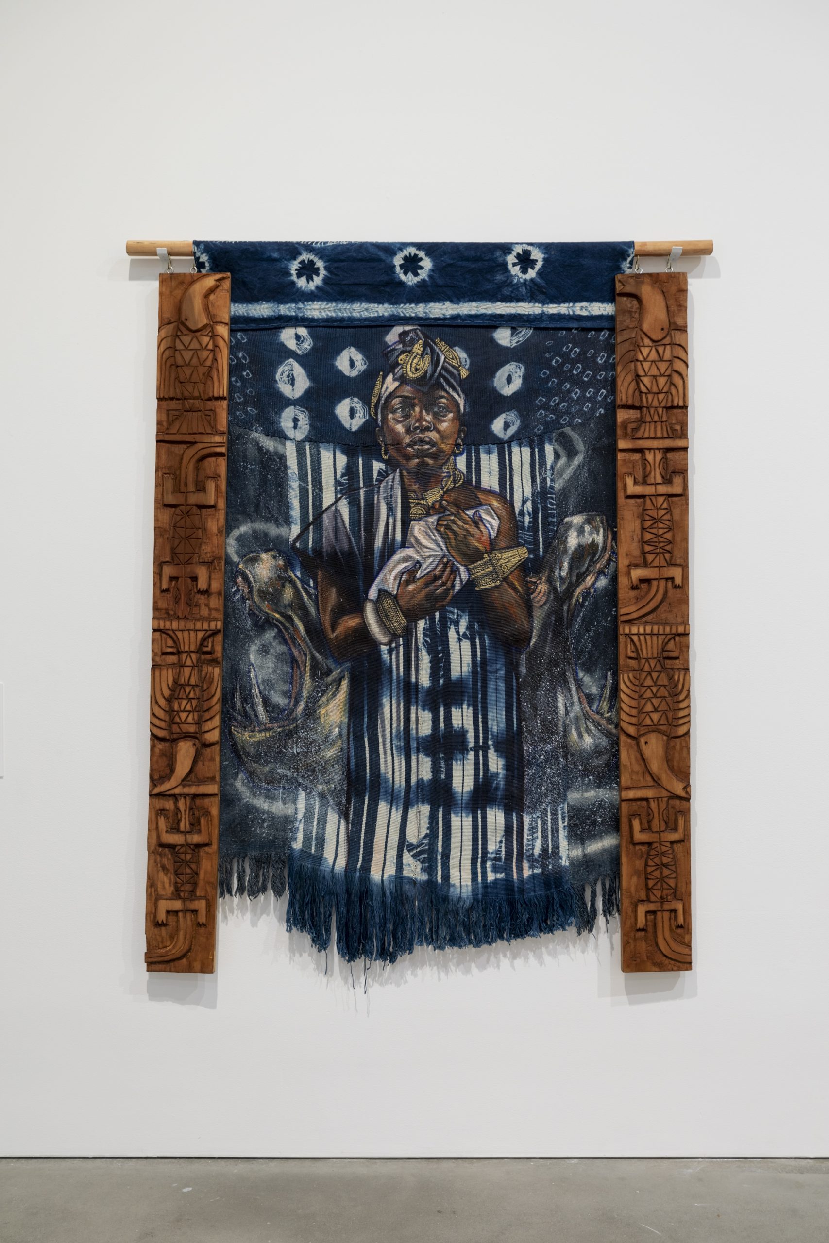 A textile artwork featuring a woman holding a baby, adorned with a blue indigo pattern. The piece is framed by ornate wooden carvings on both sides, depicting intricate designs. Several other people are subtly integrated into the background.
