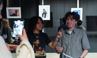 A teen interviews a another teen with a microphone, while another person records them on a smartphone. Portraits hang on the wall behind them.