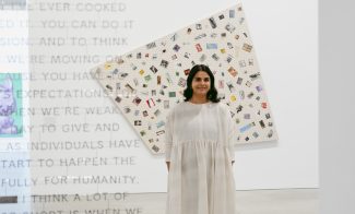 A woman in a light-colored dress stands in an art gallery, smiling. She is in front of a large, patterned artwork. Text is partially visible on a transparent panel in the foreground. The setting is modern and minimalistic.