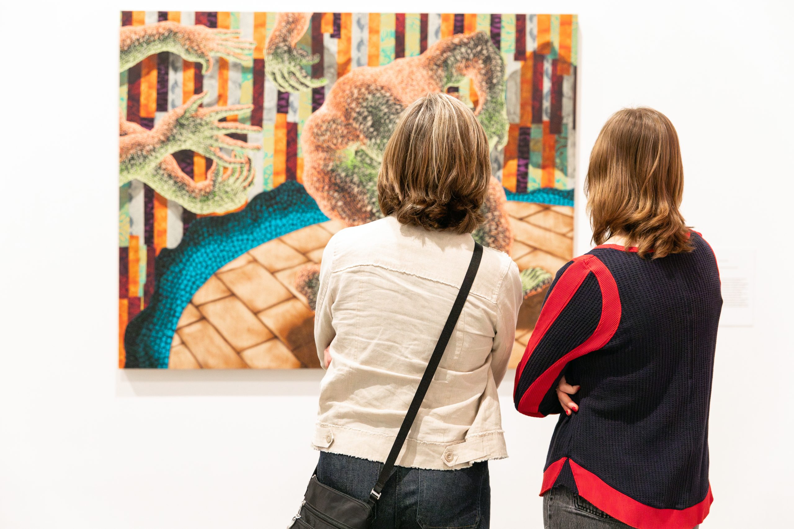 Two people with shoulder-length hair stand closely together looking at a brightly colored painting.